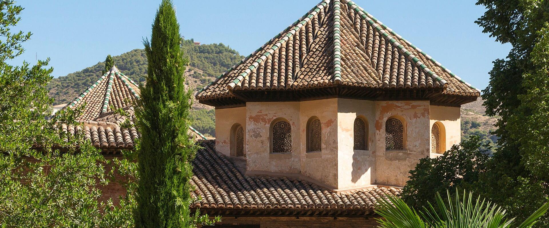 A view of the terracotta-tiled roofs and octagonal turrets of the Palace of the Comares within the Alhambra complex in Granada, Spain, framed by cypress trees and hills.