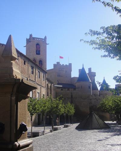 A cobblestone plaza with a stone fountain in the foreground, trees, and a medieval-style castle complex with conical-roofed towers and a flag flying in the background under a clear blue sky.