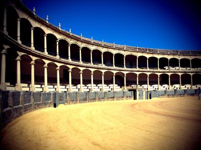 The image shows the interior of a traditional Spanish bullring with a sandy arena floor, a low stone and wood barrier, and two tiers of arched colonnades under a clear blue sky.