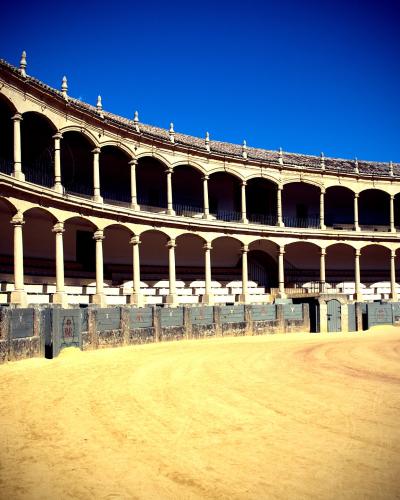 The image shows the interior of a traditional Spanish bullring with a sandy arena floor, a low stone and wood barrier, and two tiers of arched colonnades under a clear blue sky.