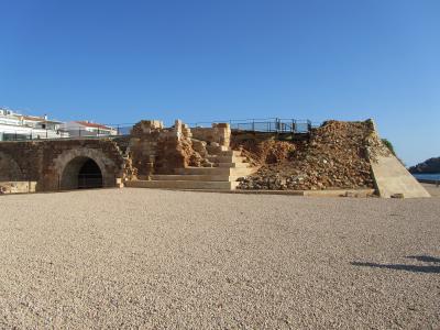 The image shows the stone ruins of a coastal fortification with an arched tunnel entrance, situated on a pebble beach under a clear blue sky, with modern white buildings visible in the background.