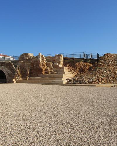 The image shows the stone ruins of a coastal fortification with an arched tunnel entrance, situated on a pebble beach under a clear blue sky, with modern white buildings visible in the background.