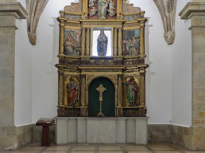 The image shows an ornate, gilded wooden altarpiece (retablo) with multiple painted panels and sculptures, situated within a stone-arched chapel or side chapel of a church.