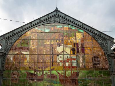 A large, ornate arched stained-glass window set within a dark metal framework, depicting historical scenes including ships and buildings, with a crest at the apex.