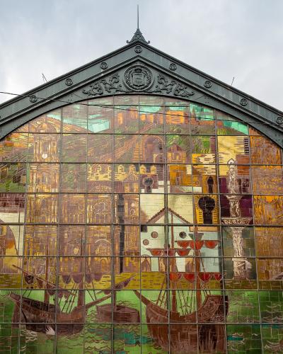 A large, ornate arched stained-glass window set within a dark metal framework, depicting historical scenes including ships and buildings, with a crest at the apex.