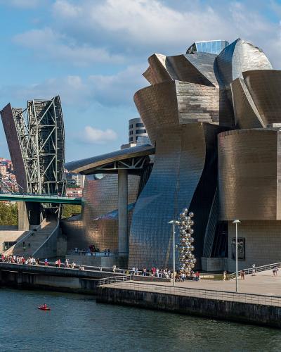 A view of the Guggenheim Museum Bilbao, a large titanium-clad building with a distinctive curvilinear design, situated on the bank of a river with a bridge and city buildings in the background.