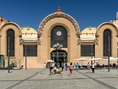 A large, ornate, tan-colored building with a distinctive arched facade, a central clock, and decorative tilework on the roof, situated in a paved public square with people and outdoor cafe seating.