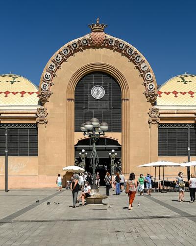 A large, ornate, tan-colored building with a distinctive arched facade, a central clock, and decorative tilework on the roof, situated in a paved public square with people and outdoor cafe seating.