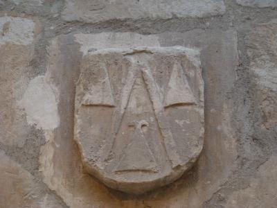 A close-up photograph of a weathered stone coat of arms carved into a rough masonry wall, featuring a shield with three conch shells (scallop shells) arranged in a triangular pattern.
