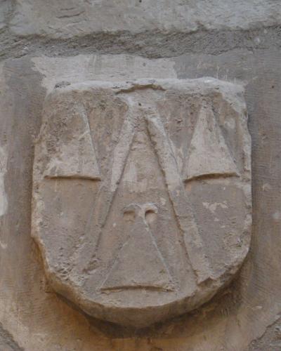 A close-up photograph of a weathered stone coat of arms carved into a rough masonry wall, featuring a shield with three conch shells (scallop shells) arranged in a triangular pattern.