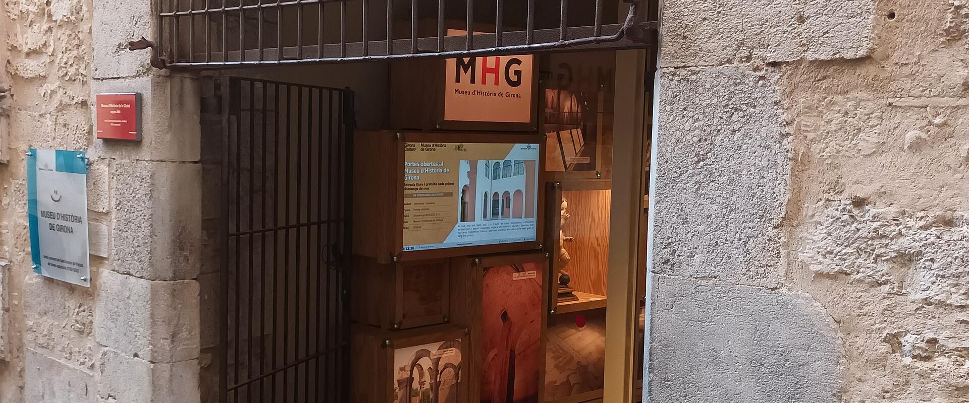 The image shows the entrance to a building with a stone facade, featuring a metal security gate, a blue and white sign on the left wall, and an interior display with a sign reading "MHG Museu d'Història de Girona".