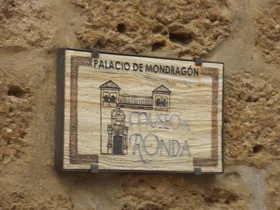 A wooden sign mounted on a rough stone wall, displaying the text "PALACIO DE MONDRAGÓN" at the top and "MUSEO DE RONDA" below an illustration of a building facade.