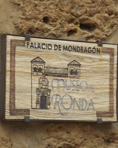A wooden sign mounted on a rough stone wall, displaying the text "PALACIO DE MONDRAGÓN" at the top and "MUSEO DE RONDA" below an illustration of a building facade.