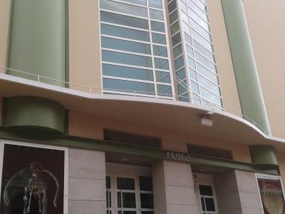 The image shows the modern exterior facade of a museum building with large glass windows, green cylindrical columns, and a poster in the lower left window displaying a dinosaur skeleton.