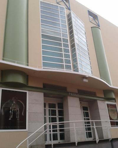 The image shows the modern exterior facade of a museum building with large glass windows, green cylindrical columns, and a poster in the lower left window displaying a dinosaur skeleton.
