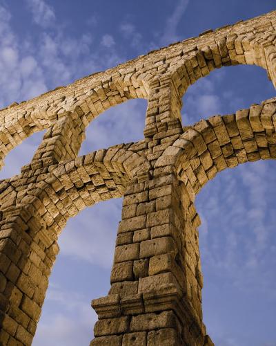 A low-angle view of a massive, two-tiered Roman stone aqueduct with large arches against a blue sky with scattered clouds.