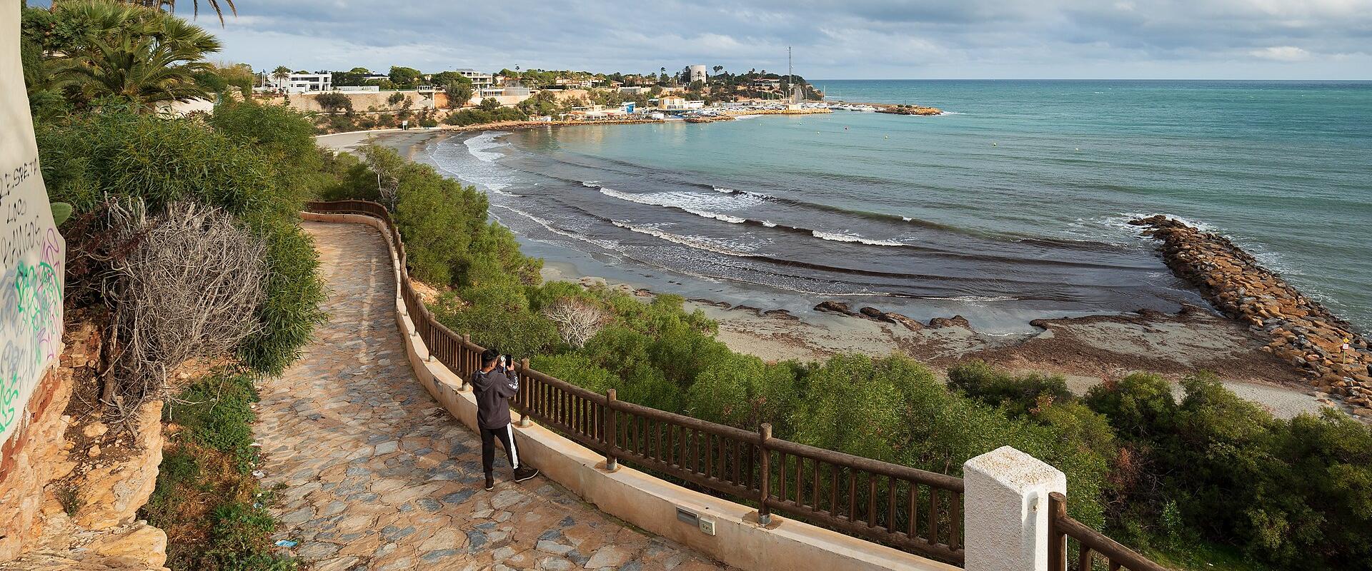 A stone-paved coastal walkway with a wooden railing overlooks a bay with a breakwater and distant buildings under a cloudy sky.