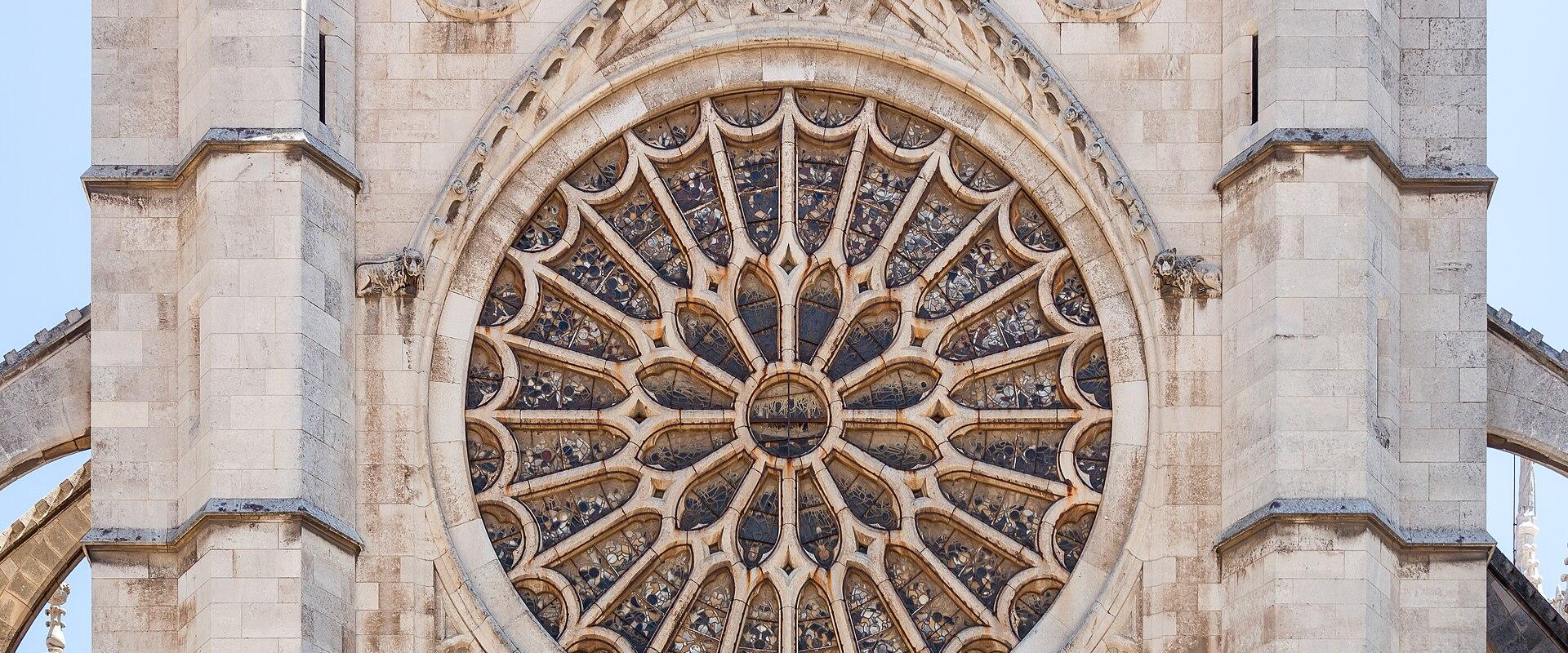 A close-up view of a large Gothic rose window set within the light-colored stone facade of a cathedral, flanked by flying buttresses.