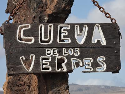 A close-up of a rustic wooden sign hanging from chains on a volcanic rock post, displaying the text "CUEVA DE LOS VERDES" in white painted letters against a backdrop of a hazy landscape.