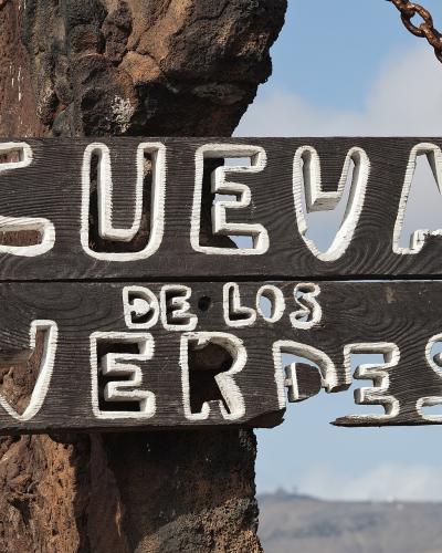 A close-up of a rustic wooden sign hanging from chains on a volcanic rock post, displaying the text "CUEVA DE LOS VERDES" in white painted letters against a backdrop of a hazy landscape.