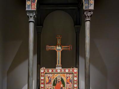 A Romanesque wooden baldachin (canopy) with painted figures and columns, enclosing a painted altar table and a processional cross, displayed in a museum setting.