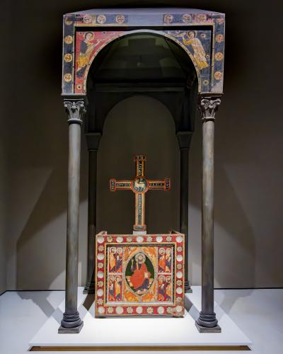 A Romanesque wooden baldachin (canopy) with painted figures and columns, enclosing a painted altar table and a processional cross, displayed in a museum setting.