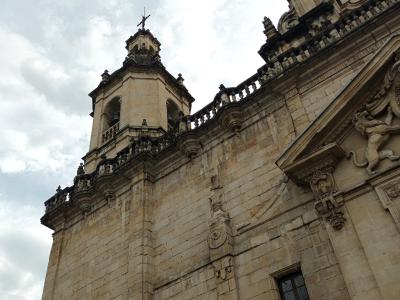 A low-angle photograph of a historic stone church facade featuring a bell tower topped with a cross and a clock tower with a visible clock face, set against a cloudy sky.