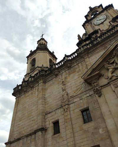 A low-angle photograph of a historic stone church facade featuring a bell tower topped with a cross and a clock tower with a visible clock face, set against a cloudy sky.