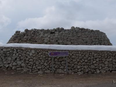 A stone talaiot (prehistoric tower) sits behind a low stone wall, with a purple directional sign in the foreground reading "Talaiot de Trebalúger".