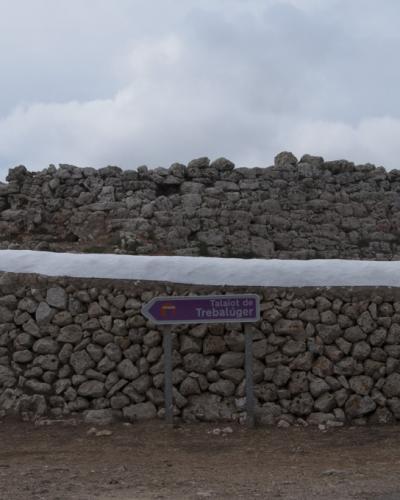 A stone talaiot (prehistoric tower) sits behind a low stone wall, with a purple directional sign in the foreground reading "Talaiot de Trebalúger".