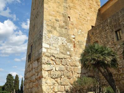 A tall, square stone tower with crenellations and corbels, part of a larger wall structure, situated next to a paved path and landscaped greenery with palm trees under a blue sky.
