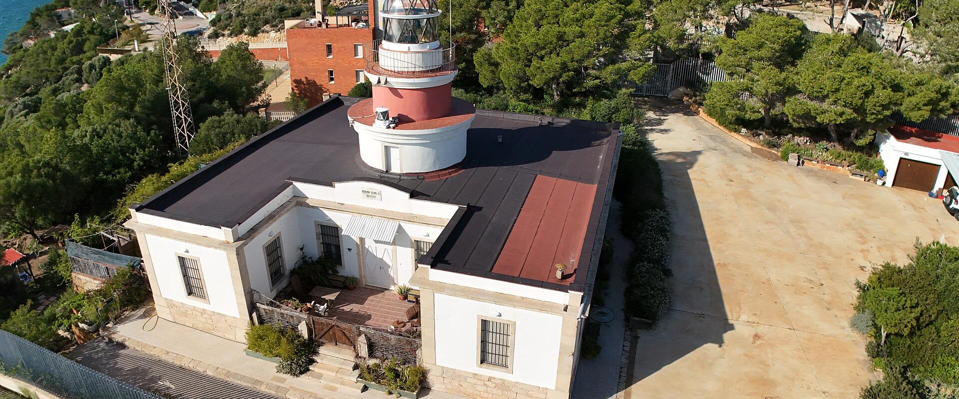 An aerial view of a white lighthouse building with a red lantern room and gallery, situated on a green hillside overlooking the sea, with residential buildings visible in the background.