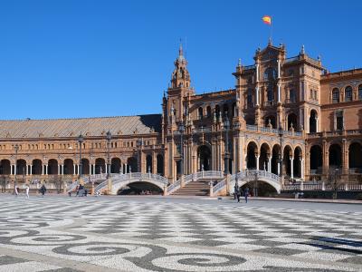 A large, semi-circular plaza with ornate Renaissance Revival architecture, tiled facades, arched colonnades, small bridges over a moat, and a distinctive black-and-white patterned pavement in the foreground, under a clear blue sky with a Spanish flag
