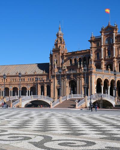 A large, semi-circular plaza with ornate Renaissance Revival architecture, tiled facades, arched colonnades, small bridges over a moat, and a distinctive black-and-white patterned pavement in the foreground, under a clear blue sky with a Spanish flag