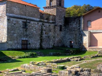 The image shows the stone ruins of a historic building complex with exposed foundation walls in a grassy courtyard, alongside a standing bell tower and adjacent structures under a clear blue sky.
