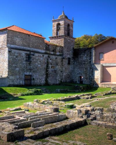 The image shows the stone ruins of a historic building complex with exposed foundation walls in a grassy courtyard, alongside a standing bell tower and adjacent structures under a clear blue sky.