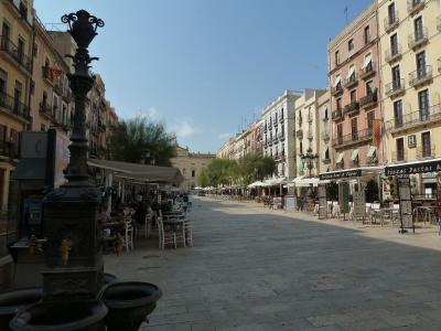 A wide, paved pedestrian plaza lined with multi-story buildings housing cafes and shops, featuring a prominent ornate black iron fountain in the foreground left and outdoor seating areas.