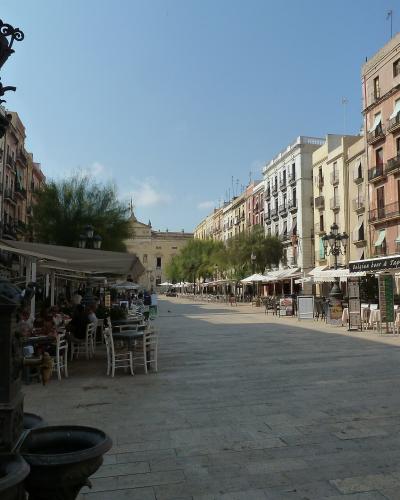 A wide, paved pedestrian plaza lined with multi-story buildings housing cafes and shops, featuring a prominent ornate black iron fountain in the foreground left and outdoor seating areas.