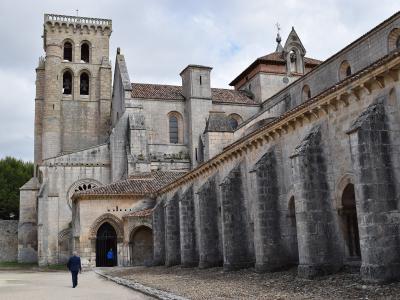 A large stone monastery complex featuring a prominent square bell tower with arched openings, a long nave with external flying buttresses, and a Gothic-style arched entrance, with a person walking on a gravel path in the foreground.