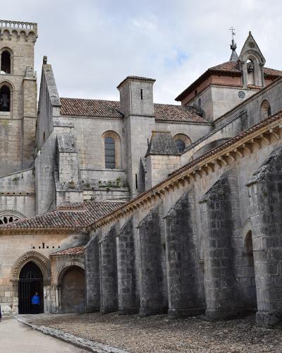 A large stone monastery complex featuring a prominent square bell tower with arched openings, a long nave with external flying buttresses, and a Gothic-style arched entrance, with a person walking on a gravel path in the foreground.
