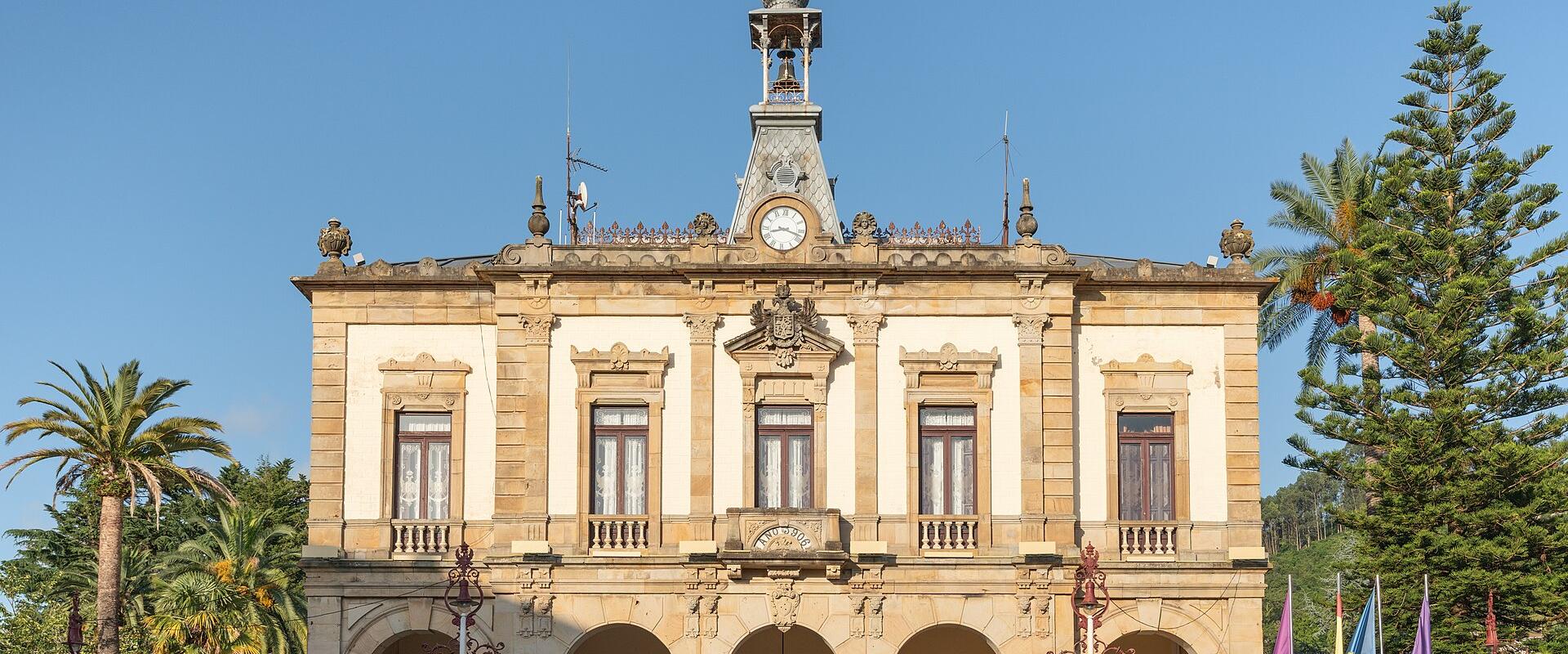 A two-story neoclassical municipal building with a central clock tower, arched ground-floor loggia, and flags flying in front, set against a clear blue sky with palm trees and other vegetation.