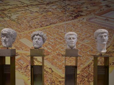 Four marble busts of Roman figures displayed on individual pedestals in front of a large wall mural depicting an ancient city plan.