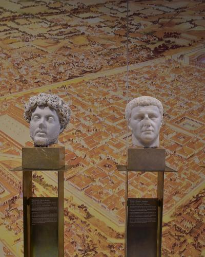 Four marble busts of Roman figures displayed on individual pedestals in front of a large wall mural depicting an ancient city plan.