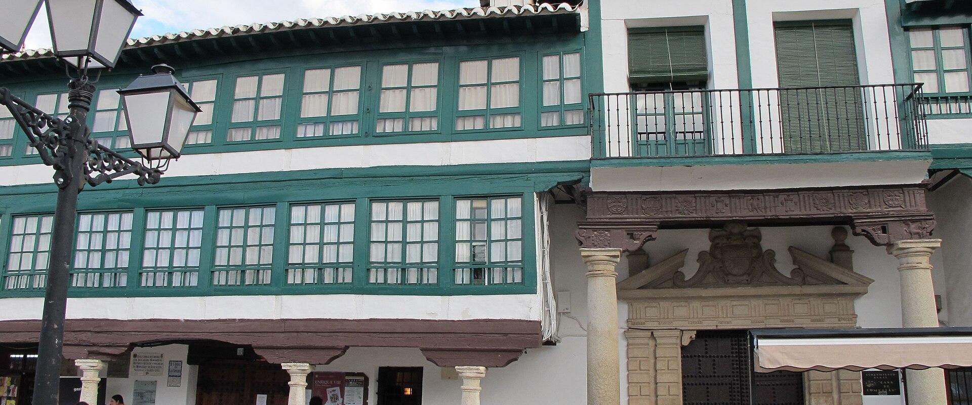 The image shows the exterior facade of a historic building with white walls, green wooden window frames and balconies, stone columns supporting an arcade, and a large ornate stone doorway, with people standing in the plaza in front.