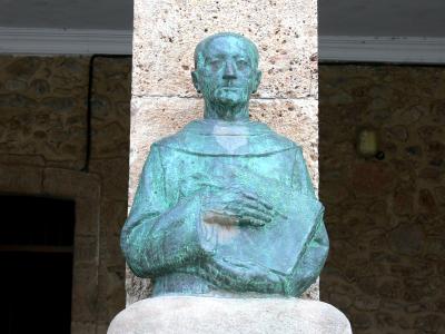A bronze bust of a man in clerical robes holding a book, set against a stone wall and column.