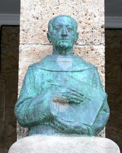 A bronze bust of a man in clerical robes holding a book, set against a stone wall and column.