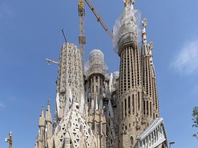 A low-angle photograph of the towering, ornate stone facade of a massive basilica under construction, featuring distinctive spires, intricate sculptural details, and large construction cranes against a clear blue sky.