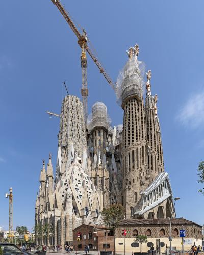 A low-angle photograph of the towering, ornate stone facade of a massive basilica under construction, featuring distinctive spires, intricate sculptural details, and large construction cranes against a clear blue sky.
