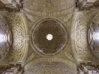 A low-angle photograph of an ornate stone vaulted ceiling featuring a central oculus, intricate Manueline-style tracery, and a coat of arms at the apex.