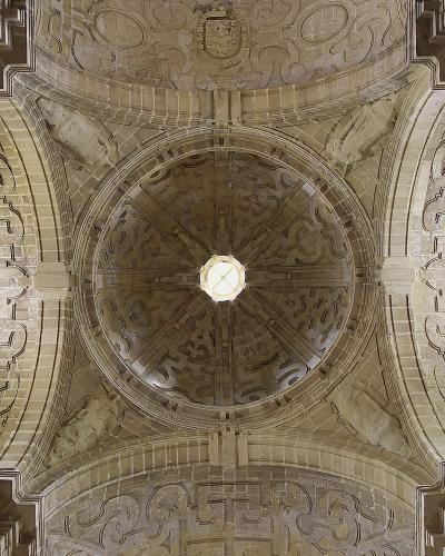 A low-angle photograph of an ornate stone vaulted ceiling featuring a central oculus, intricate Manueline-style tracery, and a coat of arms at the apex.